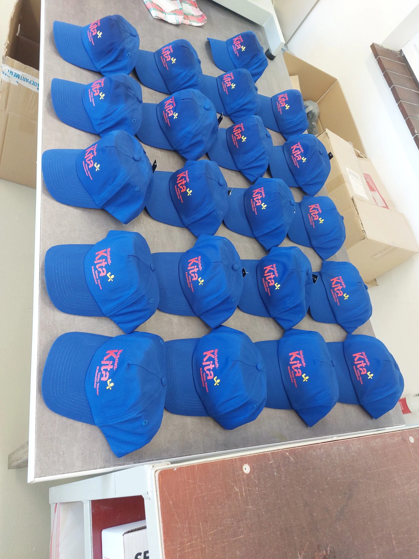 Zwanzig blaue Caps mit rotem Kita-Logo auf einem Tisch