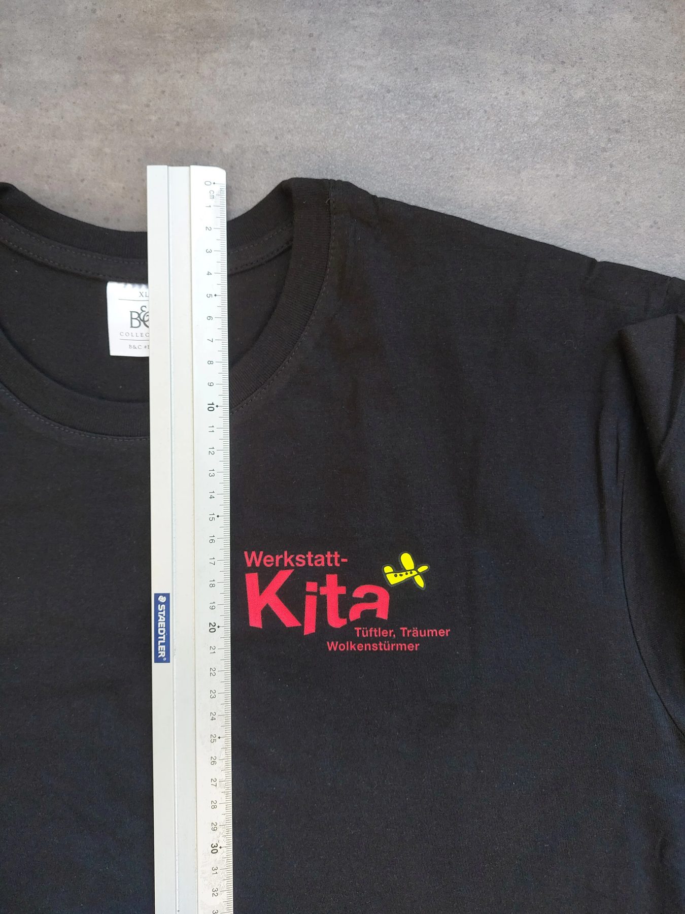 Schwarzes T-Shirt mit rotem Kita-Logo auf der Brust, daneben ein Lineal